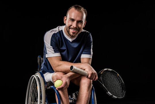 Smiling young man in sportswear sitting in wheelchair and holding tennis racquet with ball