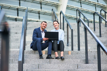 Smiling business couple with laptop. Happy people sitting on stairs. Possibilities around us.