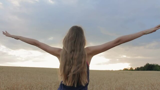 Teenage girl with raising arms and long blond hair blowing in wind enjoying nature and freedom in wheat field at sunset