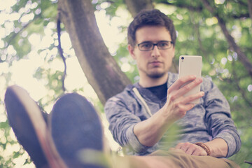 Junger Mann lehnt im Park am Baum, Blick aufs Smartphone