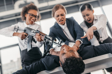 Three young angry businesswomen with rifle punishing businessman lying on table, business team...