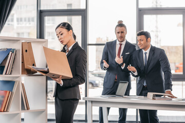 Serious young businesswoman holding folder while colleagues standing and looking behind
