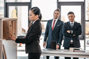 Serious young businesswoman holding folder while colleagues standing and looking behind