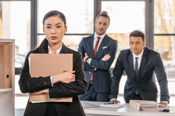 Serious young businesswoman holding folder and businessmen standing behind