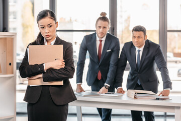 Stressed young businesswoman holding folder and businessmen standing behind