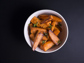 Fried sweet potatoes wedges sprinkled with wild thyme and sea salt, served in rustic grey and white bowl on black background 