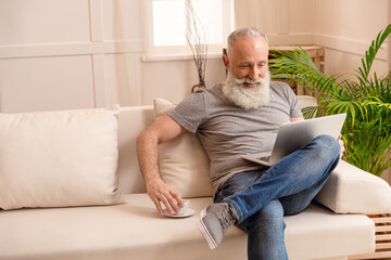 happy senior bearded man with coffee cup using laptop while sitting on sofa at home