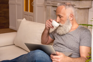 senior bearded man using laptop and drinking coffee while sitting on sofa at home