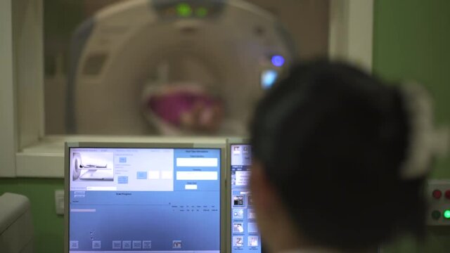 Radiology technician performs scanning in control room, crane shot, focus on the x-ray images of human body on monitor screen, shallow depth of field, patient lying on CT or MRI scanner blurred