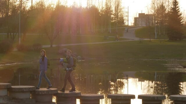 Active family with the baby passes a beautiful bridge over the river at sunset. The sun shines in the frame