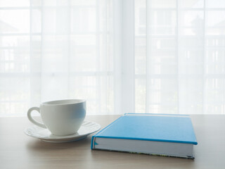 wood office table with white coffee cup and blue notebook paper on blurry beautiful white drape window texture background.