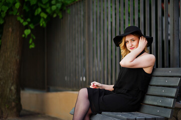 Blonde woman on black dress, necklaces and hat sitting on bench.