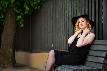 Blonde woman on black dress, necklaces and hat sitting on bench.