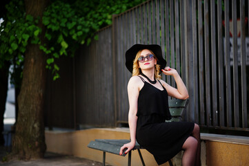 Fototapeta premium Blonde woman on black dress, sunglasses, necklaces and hat sitting on bench.