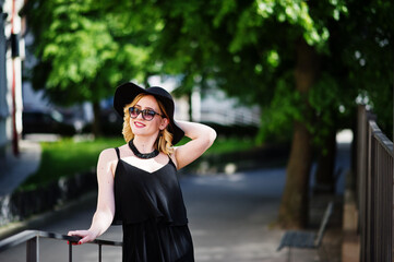 Blonde woman on black dress, sunglasses, necklaces and hat posed at streets.