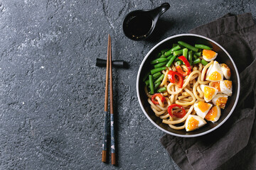 Cooking stir fry udon noodles, green beans, sliced paprika, boiled eggs, soy sauce with sesame seeds in black plate with wood chopsticks over black texture background. Flat lay. Asian style dinner
