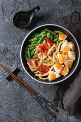 Cooking stir fry udon noodles, green beans, sliced paprika, boiled eggs, soy sauce with sesame seeds in black plate with wood chopsticks over black texture background. Flat lay. Asian style dinner