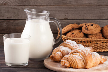 jug and glass of milk with croissants and oatmeal cookies in a wicker basket on a wooden background