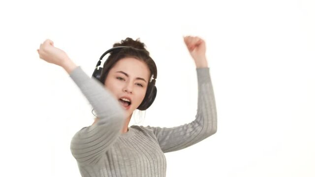 Joyful smiling Caucasian brunette girl dancing on white background listening music through black headphones on white background