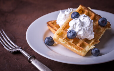 Serving belgian waffle on white plate with blueberries and double cream on rustic table.