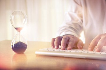 Business schedule and deadline concept. Man's hands typing on computer keyboard next to a hourglass. Concept for background, website banner, promotional materials, presentation templates, advertising.