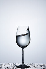 Splashing transparent water wave in the wine glass while standing on the glass with droplets against light background.
