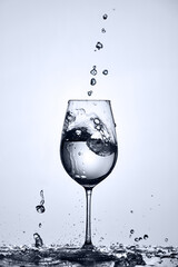 Water bubbles falling and clean water splash out of wineglass standing on the glass against light background.