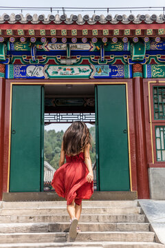Elegant woman walking through old wooden traditional chinese gate at ancient imperial temple, china summer travel. Gorgeous girl with flowing hair and fashion red dress in the wind.