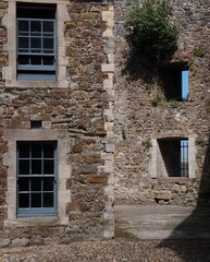 Staggered stone walls in an English courtyard with empty windows with bars and glass windows in blue frames on a sunny summer day. 