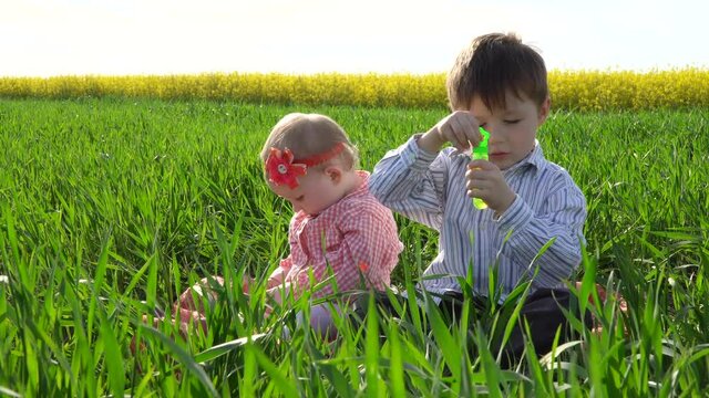 Little boy and girl are sitting in the field and blowing soap bubbles