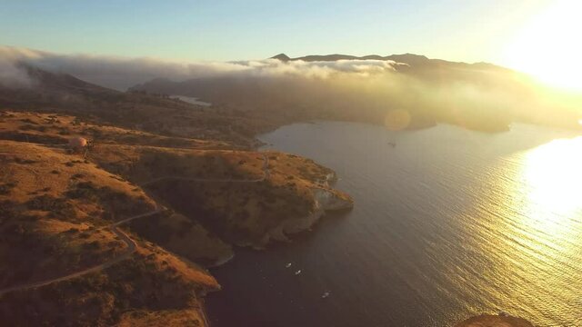 Aerial shot slowly rising, overlooking the coastline of an island at sunset. Gorgeous dazzling sunlight and cloud bank. A road winds along the cliff. sun reflects off water surface.