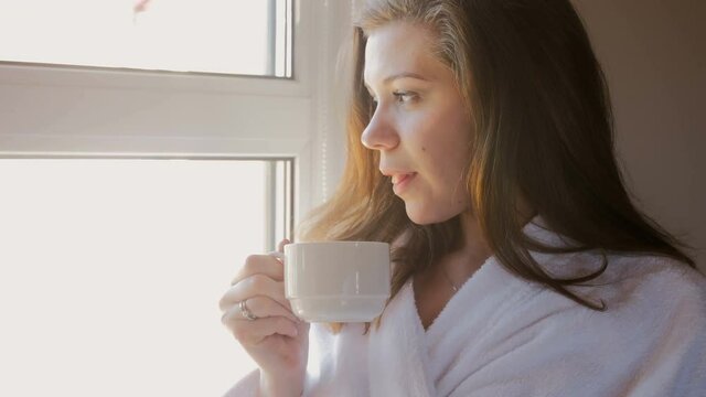 4k closeup video of beautiful young woman in bathrobe drinking coffee at window