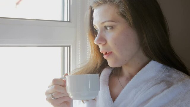Closeup video of young woman in bathrobe drinking coffee and looking out of the window
