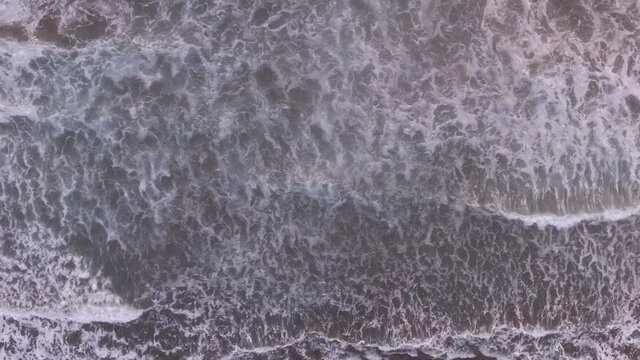 Tracking aerial top view of blue ocean waves breaking against beach. Surfers riding on boards