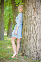 Happy American blonde woman in blue romantic chiffon dress and good figure on the beach near the river and trees