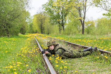 Portrait of armed woman with camouflage. Young female soldier observe with firearm on rail. Child soldier with gun in war, green grass and yellow dandelions background.  Military, army people concept