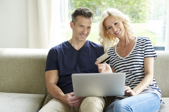 They like to shop online. Shot of a smiling middle aged couple sitting on couch and using credit card and laptop while shopping online. 