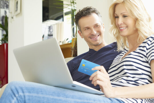 They like to shop online. Shot of a smiling middle aged couple sitting on couch and using credit card and laptop while shopping online. 