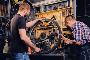Fototapeta na wymiar Two bearded mechanics fixing bicycle's wheel in a workshop.