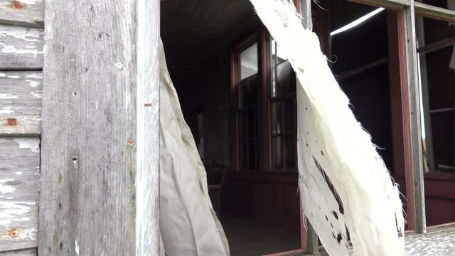 CLOSE UP Looking through the shattered window of an old ruined decaying wooden cabin. Spooky white curtain tattering in the wind. Spooky classroom with broken school decks In abandoned crumbling house