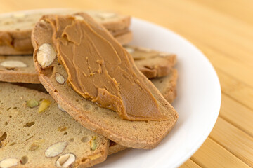 Almond pistachio crisp bread slices with cookie butter on a white plate atop a wood place mat.