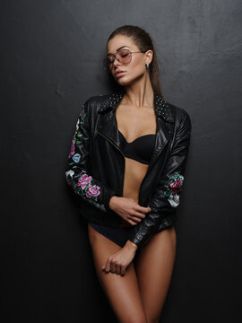Seductive, beautiful, attractive girl with sexy gorgeous tanned body is posing in the black seamless lingerie, stylish biker leather jacket and sunglasses near the dark wall  in the studio