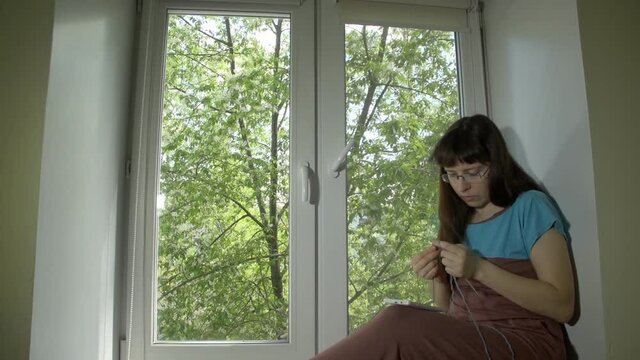 Young beautiful woman in glasses knits from blue yarn with knitting needles and uses a smartphone sitting on a windowsill at a bright spring day outside a tree with green leaves.