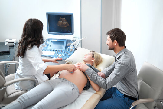 Female doctor screening stomach of woman using special tool and gel. Mom looking at picture of baby. Husband supporting and carrying about  pregnant wife on procedure of ultrasound in modern hospital.