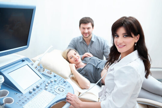 Young couple expecting of child and diagnosing in clinic. Professional female doctor using ultrasound equipment, screening of pregnant woman, smiling at camera. Doctor press buttons of special