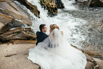 Look from behind at wedding couple sitting on the stones before a waterfall