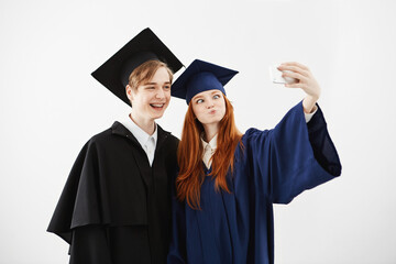 Two cheerful graduates of university fooling making selfie over white background.