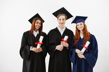 Three happy graduates smiling holding diplomas looking at camera over white background.