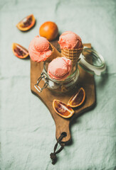 Refreshing summer blood orange ice cream or sorbet scoops in sweet waffle cones in glass jar served with fresh orange slices on rustic wooden board over light grey towel background, selective focus - obrazy, fototapety, plakaty