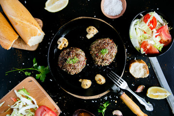 Concept of cooking hamburgers - cutlets in a cast-iron frying pan, fresh vegetables and a baguette, on a black background